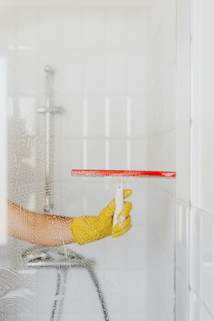 pexels-photo-4239091-4239091 A person wearing a yellow glove cleans a shower glass with a squeegee, emphasizing hygiene and cleanliness.